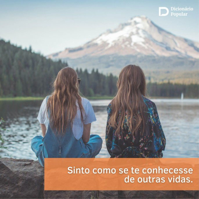 Duas garotas sentadas de costas para a foto, em um cenário com um rio a frente e montanhas com gelo no topo ao fundo