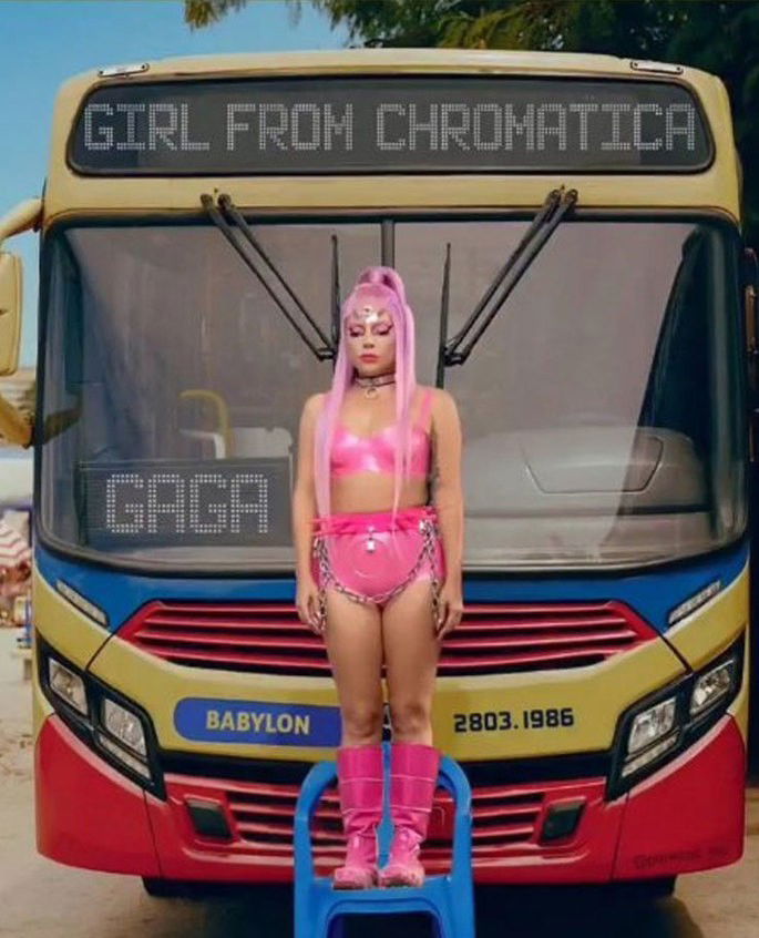Lady Gaga em pé em uma cadeira de plástico azul em frente a um ônibus