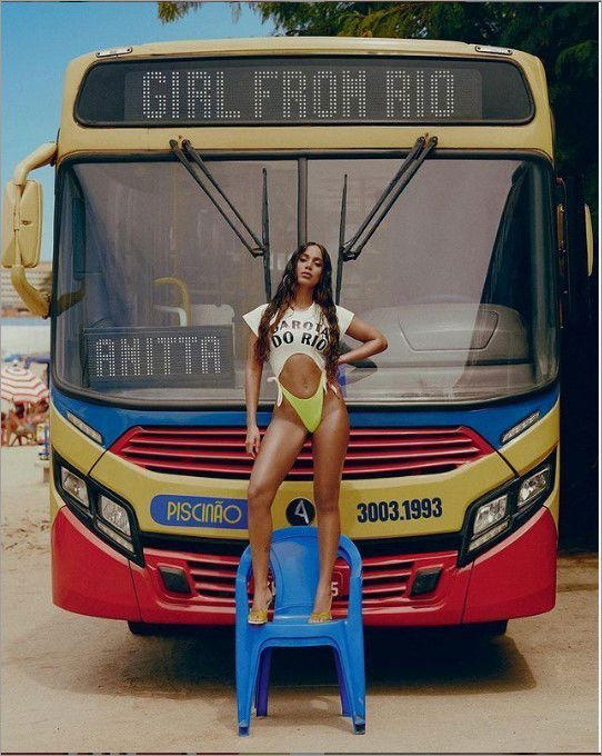 Anitta em pé em uma cadeira de plástico azul em frente a um ônibus
