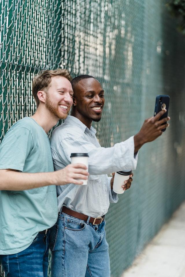 Dois homens segurando copos descatáveis de café posam para selfie em frente a uma parede de grade verde