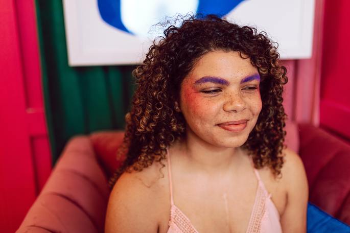 Menina com sardas e cabelo cacheado com as sobrancelhas pintadas de roxo e bochechas marcadas de blush