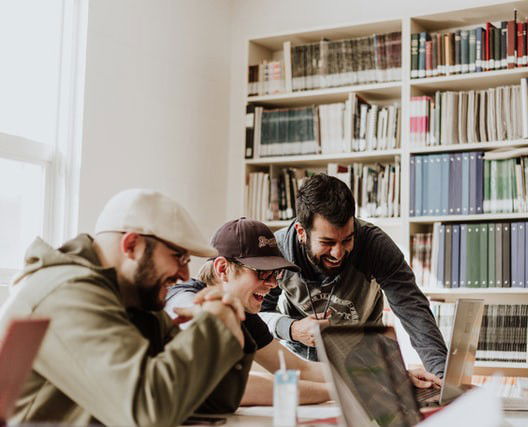 Três homens sorriem enquanto olham para a tela do computador em uma sala